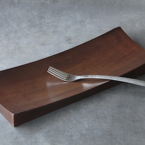 May include: A rectangular, dark brown wooden tray with a slightly concave surface. A single silver-coloured metal fork with a textured handle rests diagonally across the centre of the tray, showcasing a smooth finish and clean, simple lines.