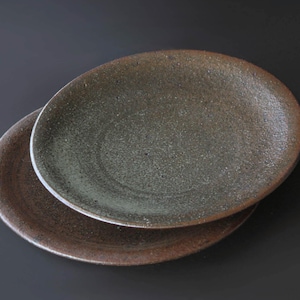 May include: Two round ceramic plates with a textured, speckled surface. The plates feature a dark brown and charcoal grey glaze with a matte finish, showing subtle variations in color and a slightly uneven, organic edge along the rim.