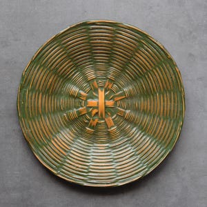 May include: A round, woven basket with a green and orange color scheme. The basket has a spiral design with a central starburst pattern. The basket is made of natural materials and is set against a gray background.