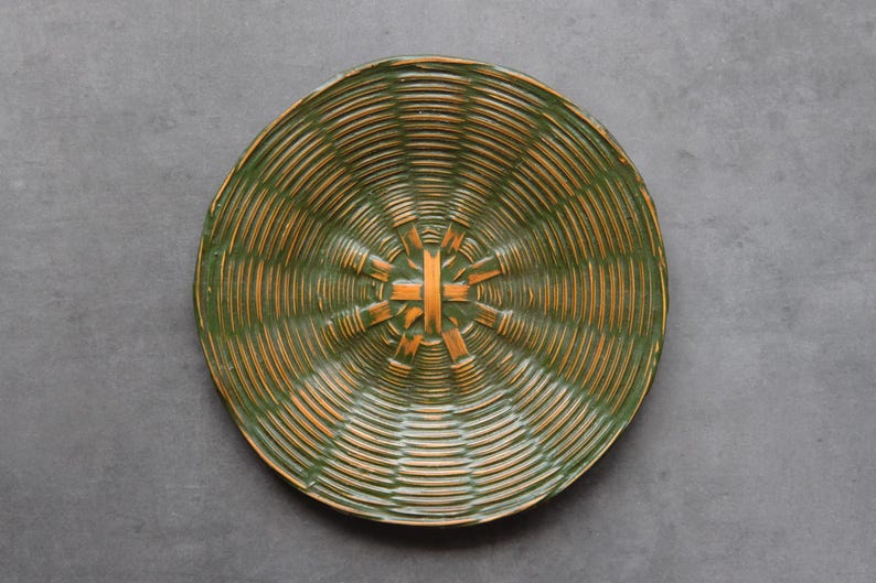 May include: A round, woven basket with a green and orange color scheme. The basket has a spiral design with a central starburst pattern. The basket is made of natural materials and is set against a gray background.