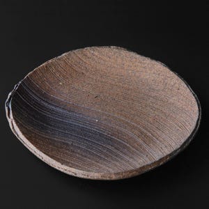 May include: A handmade, organic-shaped ceramic bowl with a textured surface. The bowl features a blend of brown and gray tones, with a dark rim and a subtle, wave-like pattern. The bowl is set against a black background.
