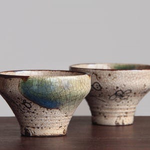 May include: Two ceramic bowls with a flared rim and a tapered base. The exterior features a cream-coloured crackle glaze finish with dark brown speckles and hand-painted black floral line art. One bowl displays a prominent blue and green splash.
