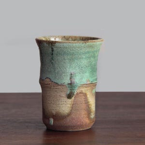 Puede incluir: Un vaso de cerámica hecho a mano con una superficie texturizada. La mitad superior es turquesa moteado, que transiciona a una base marrón cálido. La taza es cilíndrica con un borde ligeramente ensanchado.