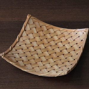 May include: A woven bamboo tray with a curved shape. The tray is made of light brown bamboo strips woven together in a basket weave pattern. The edges are neatly finished with a thin bamboo border. The tray sits on a dark brown wooden surface.