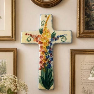 May include: A ceramic wall cross featuring a hand-painted floral design with vibrant yellow, orange, and blue blossoms extending from green leaves. The cross has a cream-colored background, a glossy finish, and a small circular hole at the top for hanging.