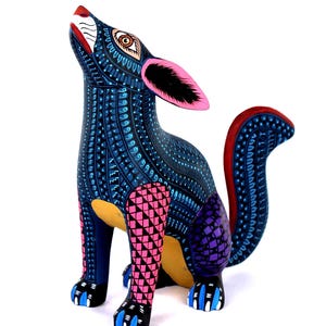 May include: A hand-painted wooden coyote figurine featuring a dark blue base color with intricate light blue, pink, and purple geometric patterns. The coyote is in a seated position with its head tilted upward, showcasing pink ears and a curved tail.