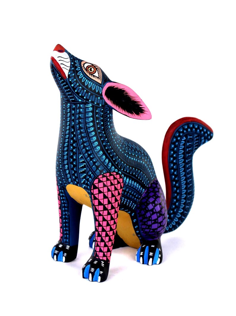 May include: A hand-painted wooden coyote figurine featuring a dark blue base color with intricate light blue, pink, and purple geometric patterns. The coyote is in a seated position with its head tilted upward, showcasing pink ears and a curved tail.