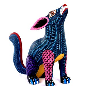 May include: A hand-painted wooden coyote figurine featuring a dark blue base color with intricate light blue, pink, and purple geometric patterns. The coyote is depicted in a howling pose with large pink ears, a yellow underbelly, and blue claws.