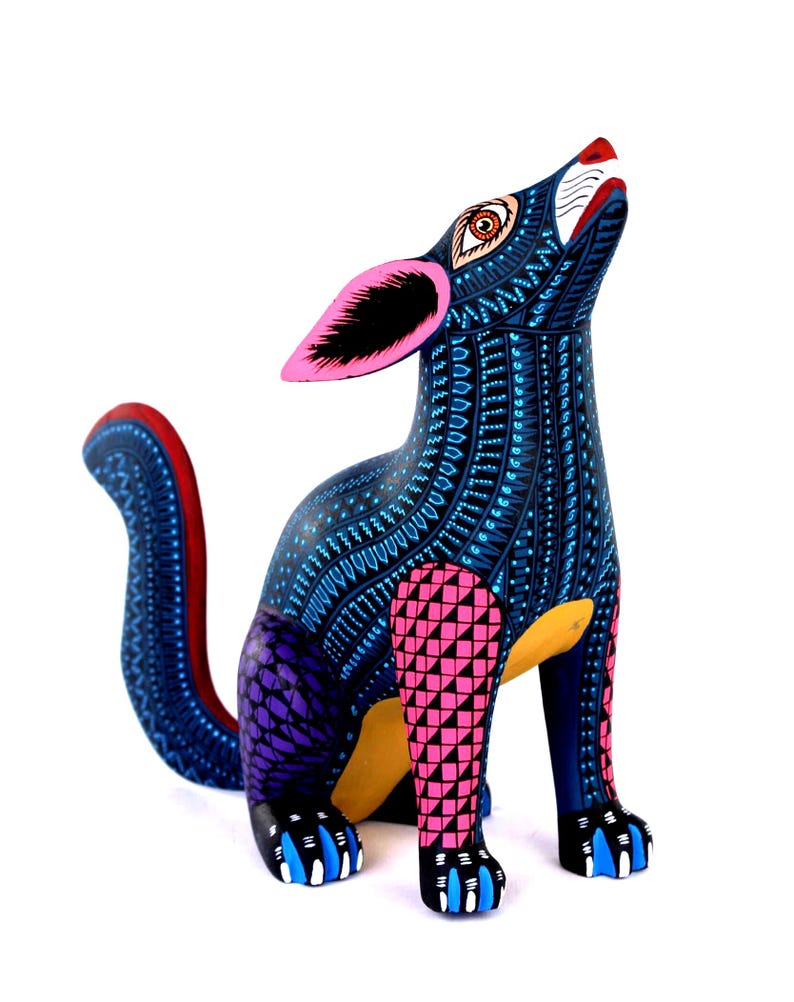 May include: A hand-painted wooden coyote figurine featuring a dark blue base color with intricate light blue, pink, and purple geometric patterns. The coyote is depicted in a howling pose with large pink ears, a yellow underbelly, and blue claws.