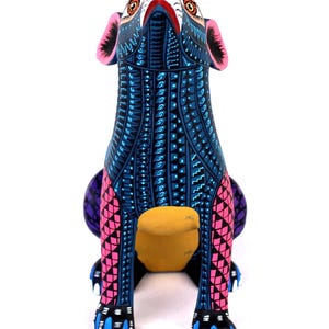 May include: A hand-painted wooden animal figurine featuring a dog-like form with a vibrant blue base color. The surface is covered in intricate geometric patterns in black, white, and pink. It has large ears, pointed eyes, and blue-tipped paws.