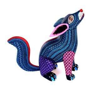 May include: A hand-painted wooden coyote figurine in a howling pose. The body is covered in intricate blue patterns with purple and pink geometric patches on the legs. It features large pink-lined ears, a red-trimmed tail, and black paws.