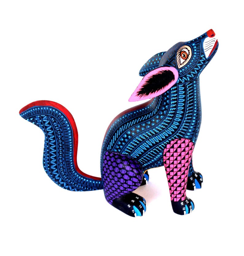 May include: A hand-painted wooden coyote figurine in a howling pose. The body is covered in intricate blue patterns with purple and pink geometric patches on the legs. It features large pink-lined ears, a red-trimmed tail, and black paws.