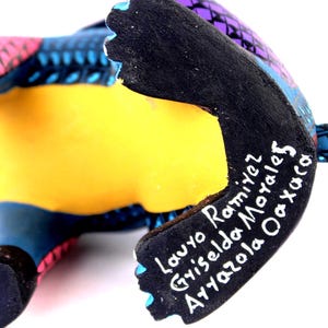 May include: A hand-painted wooden figurine featuring a vibrant purple, blue, and pink geometric pattern. The underside is painted solid black with white hand-painted text reading Lauro Ramirez, Griselda Morales, Arrazola Oaxaca.
