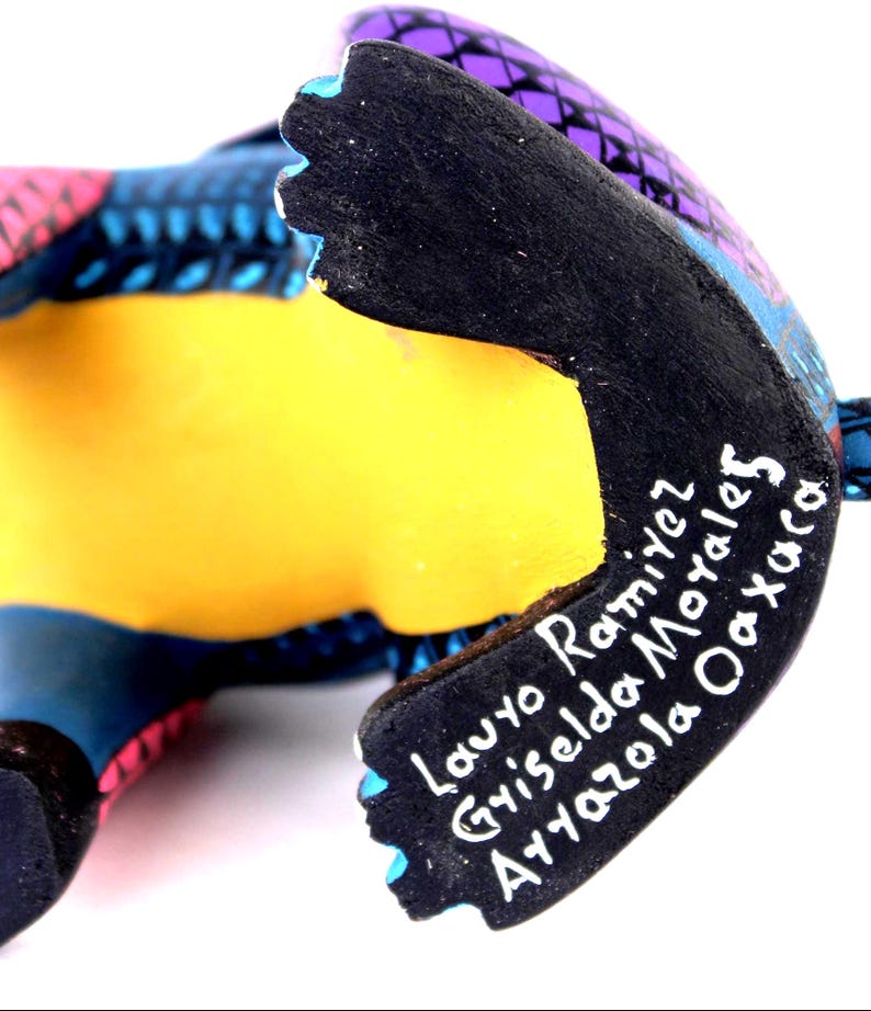 May include: A hand-painted wooden figurine featuring a vibrant purple, blue, and pink geometric pattern. The underside is painted solid black with white hand-painted text reading Lauro Ramirez, Griselda Morales, Arrazola Oaxaca.
