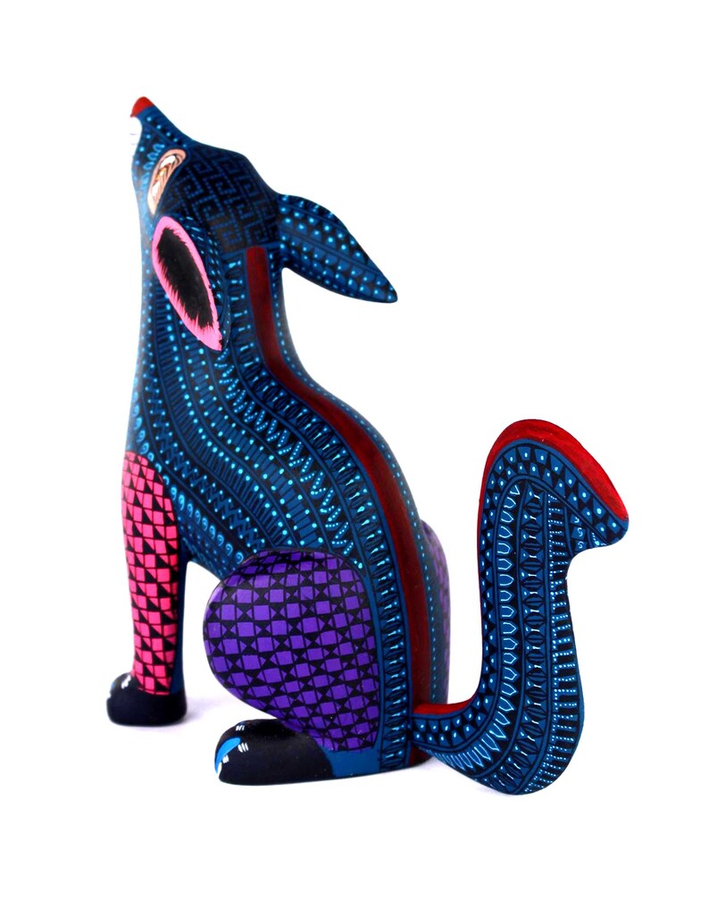 May include: A hand-painted wooden coyote figurine in a howling pose, featuring a deep blue base color with intricate geometric patterns in light blue, pink, purple, and red. The animal has pointed ears, a curved tail, and detailed dot work.