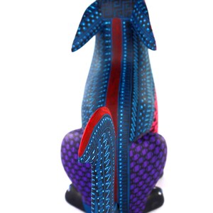 May include: A hand-painted wooden dog figurine featuring a vibrant blue base color with intricate black geometric patterns, purple textured hindquarters, and bright red accents on the ears, tail, and a central stripe along the back.