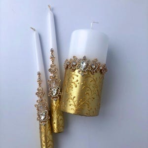 Puede incluir: Tres velas blancas con adornos dorados y de joyas. Dos son altas y delgadas, mientras que la tercera es una vela de pilar más ancha. La vela de pilar tiene un diseño floral dorado y una banda de joyas. Las otras velas tienen detalles de joyas.