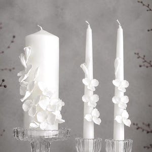Puede incluir: Una vela de pilar blanca grande y dos velas cónicas blancas delgadas, cada una decorada con delicadas aplicaciones florales blancas. Las velas se exhiben en candelabros de vidrio transparente acanalado, mostrando una estética limpia y monocromática.