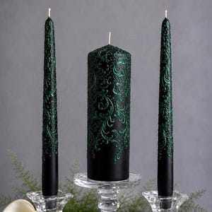 Könnte beinhalten: Ein Set aus drei schwarzen Wachskerzen mit erhabenen, metallisch grünen Filigranmustern. Zwei hohe, schlanke Stabkerzen flankieren eine einzelne, breitere zylindrische Stumpenkerze. Jede Kerze steht in einem klaren Kerzenhalter aus Glas.