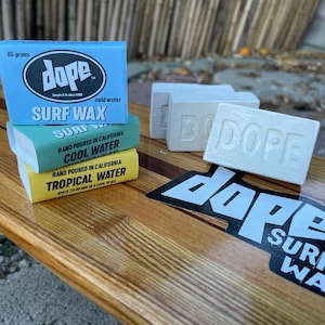 May include: Three bars of Dope surf wax stacked on top of each other. The top bar is blue and says "Dope Surf Wax" in white text. The middle bar is green and says "Hand Poured in California Cool Water" in white text. The bottom bar is yellow and says "Hand Poured in California Tropical Water" in white text. There is a black and white Dope Surf Wax sticker on a wooden surfboard.
