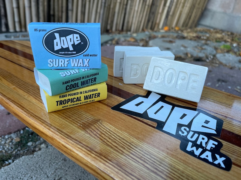 May include: Three bars of Dope surf wax stacked on top of each other. The top bar is blue and says "Dope Surf Wax" in white text. The middle bar is green and says "Hand Poured in California Cool Water" in white text. The bottom bar is yellow and says "Hand Poured in California Tropical Water" in white text. There is a black and white Dope Surf Wax sticker on a wooden surfboard.