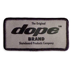Peut inclure: Écusson rectangulaire avec le texte "dope BRAND" en noir, avec "The Original" au-dessus et "Skateboard Products Company" en dessous. L'écusson a un fond gris et une bordure violet foncé.