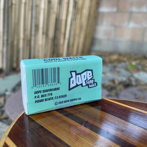 May include: A teal blue bar of surf wax with the text "dope surf wax" in white. The bar is labelled with the text "Cool Water" and has a barcode and the text "Dope Surfboards, P.O. Box 774, Pismo Beach, CA 93420, 2021 Dope Supply Co."