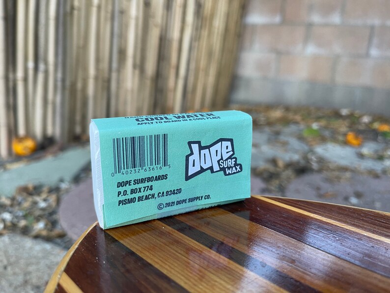 May include: A teal blue bar of surf wax with the text "dope surf wax" in white. The bar is labelled with the text "Cool Water" and has a barcode and the text "Dope Surfboards, P.O. Box 774, Pismo Beach, CA 93420, 2021 Dope Supply Co."