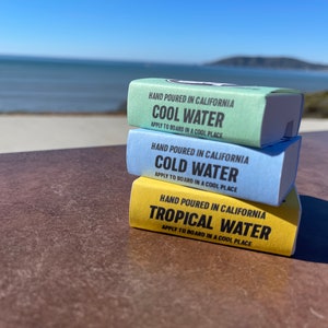May include: Three bars of surfboard wax stacked on top of each other. The top bar is yellow and says "HAND POURED IN CALIFORNIA TROPICAL WATER APPLY TO BOARD IN A COOL PLACE". The middle bar is blue and says "HAND POURED IN CALIFORNIA COLD WATER APPLY TO BOARD IN A COOL PLACE". The bottom bar is green and says "HAND POURED IN CALIFORNIA COOL WATER APPLY TO BOARD IN A COOL PLACE".