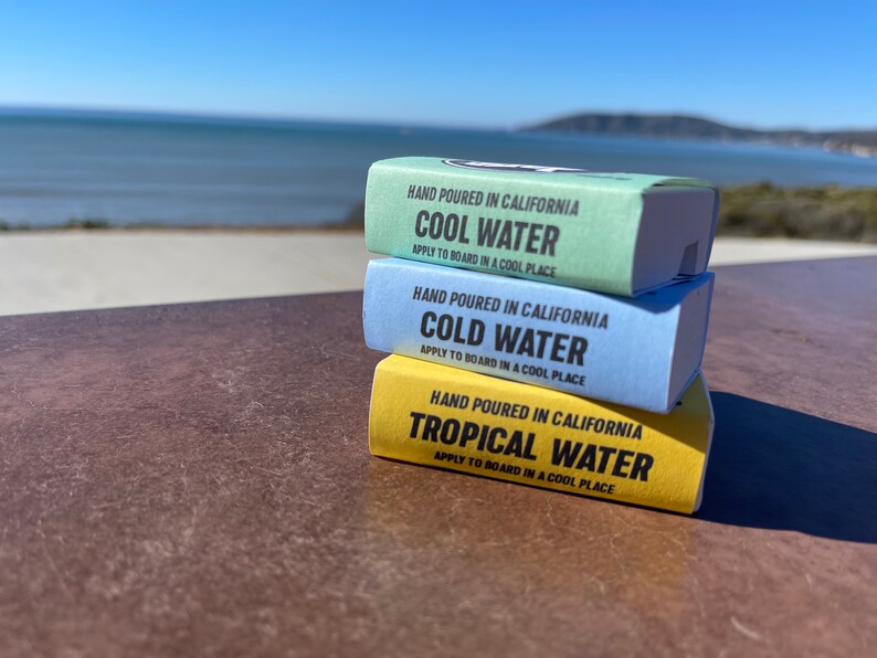 May include: Three bars of surfboard wax stacked on top of each other. The top bar is yellow and says "HAND POURED IN CALIFORNIA TROPICAL WATER APPLY TO BOARD IN A COOL PLACE". The middle bar is blue and says "HAND POURED IN CALIFORNIA COLD WATER APPLY TO BOARD IN A COOL PLACE". The bottom bar is green and says "HAND POURED IN CALIFORNIA COOL WATER APPLY TO BOARD IN A COOL PLACE".