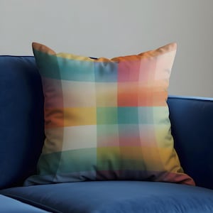 Peut inclure: Un coussin carré décoratif avec un motif à carreaux coloré. Le coussin présente un quadrillage de lignes entrecroisées dans des tons de turquoise, jaune, orange, rose et blanc. Il est posé sur un canapé bleu.