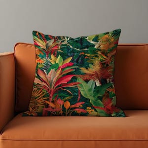 Könnte beinhalten: Quadratisches Kissen mit einem lebendigen tropischen Dschungel-Print. Das Kissen zeigt üppiges grünes, rotes, orangefarbenes und gelbes Laub. Das Kissen liegt auf einem orangefarbenen Sofa.