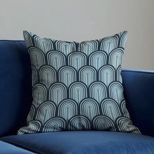Könnte beinhalten: Quadratisches Zierkissen mit einem sich wiederholenden Bogenmuster in Blau- und Weißtönen. Das Kissen liegt auf einem blauen Sofa und erzeugt ein harmonisches Farbschema. Das Design erinnert an den Art-Deco-Stil.