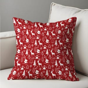 May include: A red decorative pillow with a repeating white pattern of Christmas trees, reindeer, houses, and snowmen. The pillow is square and rests on a white sofa, perfect for holiday decor.
