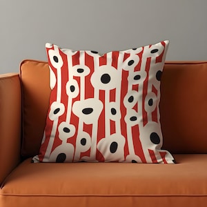 May include: A decorative throw pillow with a bold, abstract design. The pillow features a pattern of vertical red stripes with white, elongated shapes and black circular accents. The pillow is placed on an orange sofa.