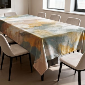 Puede incluir: Mantel rectangular con un diseño abstracto de acuarela en tonos amarillos, azules y marrones. El mantel está colocado sobre una mesa de comedor, rodeado de sillas beige con patas negras. El tema general es la decoración del hogar.