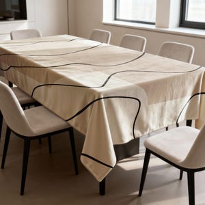 Peut inclure: Une nappe rectangulaire beige avec un motif abstrait noir recouvre une table à manger. La nappe présente un motif géométrique avec des lignes noires incurvées. La table est entourée de chaises beiges avec des pieds noirs.