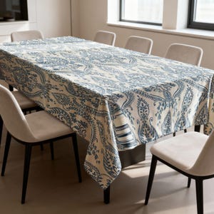 May include: A cream-coloured rectangular tablecloth featuring a blue paisley design. The cloth is spread over a large dining table, surrounded by chairs. The intricate pattern is consistent across the entire surface of the tablecloth.