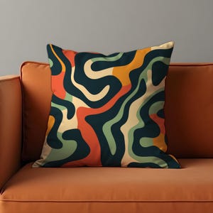May include: Square cushion with a retro abstract design. The pattern has flowing, organic shapes in navy, orange, green, and cream. The cushion is on an orange sofa, creating a vibrant contrast. The cushion is approximately 40cm x 40cm.