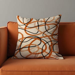 May include: Square throw pillow with a cream background and an abstract design of intertwined orange and black lines. The pillow is placed on an orange sofa, creating a complementary color scheme.