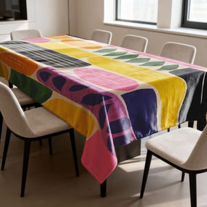 May include: A rectangular tablecloth with a vibrant abstract design. The pattern includes large, irregular shapes in yellow, pink, orange, green, blue, and black on a cream backdrop. The tablecloth covers a dining table with white chairs.