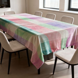 Puede incluir: Un mantel rectangular con un patrón geométrico colorido. El diseño presenta grandes rectángulos curvos en tonos de rosa, verde, morado y blanco. El mantel está colocado sobre una mesa de comedor, rodeado de sillas de tonos neutros.