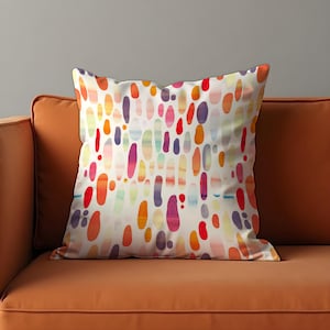 Könnte beinhalten: Ein quadratisches Zierkissen mit weißem Hintergrund und einem farbenfrohen Muster aus vertikalen ovalen Formen in Rot-, Orange-, Gelb-, Lila- und Blautönen. Das Kissen liegt auf einem orangefarbenen Sofa.