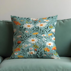 May include: Square throw pillow with a floral pattern. The pillow features a light blue background with a repeating pattern of white daisies, orange and yellow flowers, and green leaves. The pillow is placed on a teal couch.