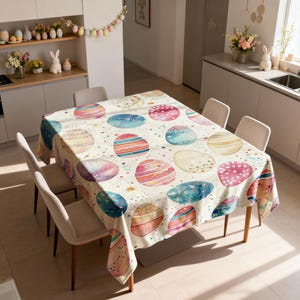 May include: A rectangular Easter tablecloth with a repeating pattern of colourful, watercolour-style Easter eggs in shades of blue, pink, yellow, and orange. The eggs are scattered across a cream background with small, speckled dots. The tablecloth is draped over a wooden table.