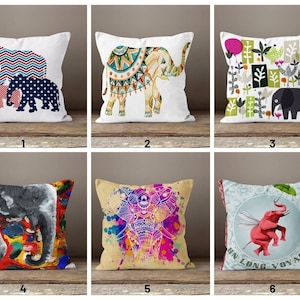 May include: Six different cushion designs featuring elephants. The cushions are all square and have a white background. The elephants are all different colours and patterns. The first cushion has a family of elephants with red, white, and blue patterns. The second cushion has a single elephant with a colourful mandala pattern. The third cushion has a single elephant with a green, pink, and white pattern. The fourth cushion has a single elephant with a colourful abstract pattern. The fifth cushion has a single elephant with a pink and purple watercolour pattern. The sixth cushion has a single elephant with a pink and white pattern and the text "En Long Voyage".