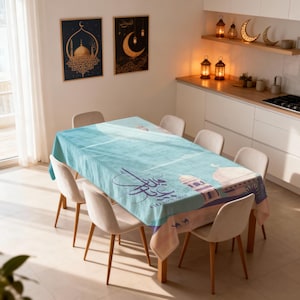 May include: A turquoise tablecloth featuring Arabic calligraphy and mosque designs, draped over a wooden table surrounded by beige chairs. Two framed artworks adorn the wall, and the kitchen is decorated with lanterns.