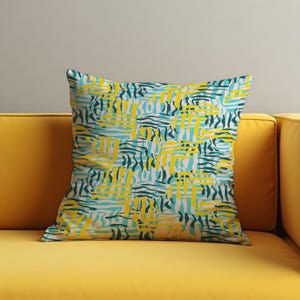 May include: Square throw pillow with a geometric pattern in yellow, teal, and dark green on a cream background. The pillow is placed on a yellow sofa. The pattern consists of intersecting lines and shapes.