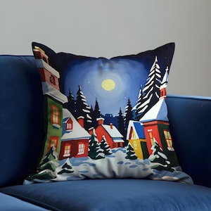 Puede incluir: Cojín decorativo con una escena de un pueblo invernal bajo la luna llena. El diseño incluye casas cubiertas de nieve en rojo, verde y amarillo, rodeadas de árboles de hoja perenne. El cojín está sobre un sofá azul.