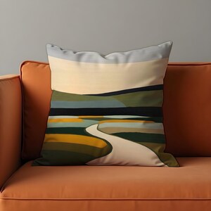 May include: Decorative throw pillow with an abstract landscape design. The pillow features a winding path through fields of green, yellow, and blue, under a pale yellow sky. The pillow is resting on an orange sofa.